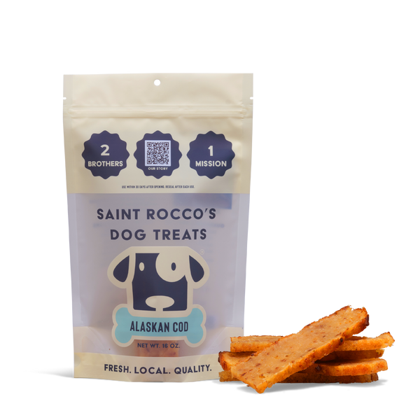 A bag labeled Alaskan Cod Dog Treats stands upright beside several rectangular human-grade treats. The packaging highlights 2 brothers, 1 mission, and emphasizes fresh, local, quality.