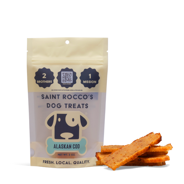 A package of Alaskan Cod dog treats stands upright beside several treat pieces. The resealable bag features a dog face illustration and highlights its quality, mission, and delicious human-grade ingredients.