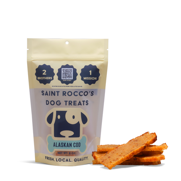 A package of Alaskan Cod dog treats stands upright beside several rectangular sticks. The packaging shows a cartoon dog and notes the treats are fresh, high-quality, and delicious human-grade snacks for dogs.